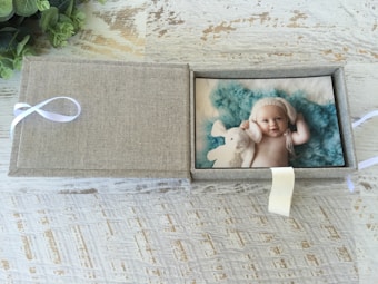 A textured box with a light brown fabric exterior is open to reveal a photo of a baby inside. The baby is smiling, wearing a white knitted hat, and cuddling a white stuffed animal against a fluffy teal backdrop. The box is placed on a rustic wooden surface and features a white ribbon on the side.