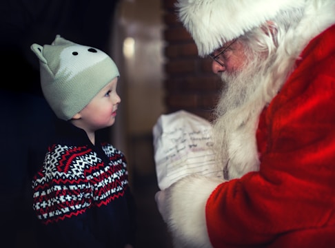 A child wearing a winter hat and a festive sweater stands facing a figure dressed as Santa Claus. The child has a look of wonder and excitement, while Santa Claus holds a piece of paper that appears to be a wish list.