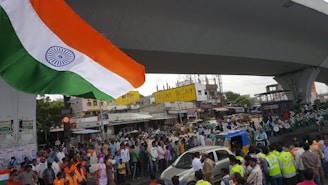 A large crowd gathered under an elevated roadway in an urban area. At the forefront, the Indian national flag is prominently displayed. The crowd includes a mix of people, some in uniform or reflective vests, and vehicles like cars and auto-rickshaws. Buildings and shops are visible in the background.