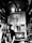 A black and white image depicts a coffee shop interior with a barista behind the counter. The focus is on the vintage espresso machine and a neon sign reading 'CAFÉ' with a steaming coffee cup icon. The setting has a cozy, rustic feel with wooden accents and shelves displaying cups.