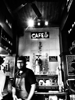 A black and white image depicts a coffee shop interior with a barista behind the counter. The focus is on the vintage espresso machine and a neon sign reading 'CAF&Eacute;' with a steaming coffee cup icon. The setting has a cozy, rustic feel with wooden accents and shelves displaying cups.