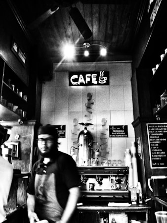 A black and white image depicts a coffee shop interior with a barista behind the counter. The focus is on the vintage espresso machine and a neon sign reading 'CAF&Eacute;' with a steaming coffee cup icon. The setting has a cozy, rustic feel with wooden accents and shelves displaying cups.