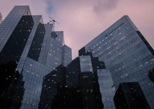 Tall modern skyscrapers with reflective glass facades rise against a cloudy sky. The buildings are structured with multiple panels, creating a geometric pattern with high transparency. Sparse lighting inside suggests the buildings are occupied.