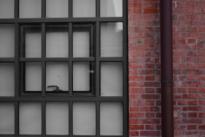 A window with multiple square panes is set against a red brick wall. The window frame is black, and there is a small handle visible on the lower pane. A vertical pipe runs parallel to the window on the right side.