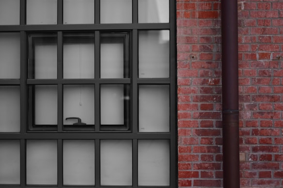 A window with multiple square panes is set against a red brick wall. The window frame is black, and there is a small handle visible on the lower pane. A vertical pipe runs parallel to the window on the right side.