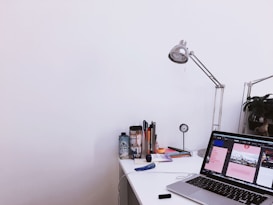 A tidy workspace featuring a laptop open on a desk, displaying a design software interface. On the desk are various office supplies, including pens, a water bottle, a lamp, and a clock. A plant is visible in the background, adding a touch of greenery.