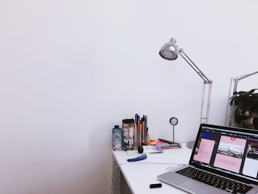 A tidy workspace featuring a laptop open on a desk, displaying a design software interface. On the desk are various office supplies, including pens, a water bottle, a lamp, and a clock. A plant is visible in the background, adding a touch of greenery.