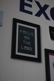 A framed poster with the text 'FOCUS ON THE LORD' hangs on a textured wall. The text is slightly blurred, and the frame is simple with a black border. Part of a large blue letter 'E' from another piece of text is visible above the frame, while another framed item with unreadable content is partially visible to the right.