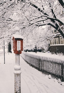 A snow-covered street with a red emergency box in the foreground. The trees and houses are blanketed in snow, creating a winter wonderland atmosphere. A wooden fence lines the sidewalk, and snow tracks are visible on the path.