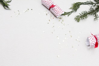Gift crackers wrapped in white paper with red dots and tied with red ribbons are scattered among green pine branches. Shiny, metallic confetti is sprinkled on a light-colored surface.