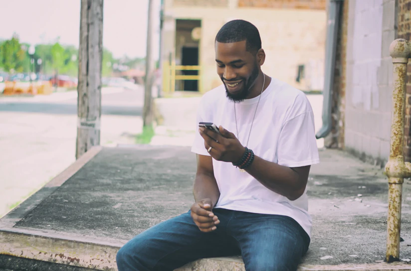 A person is sitting on a concrete platform near a building, casually dressed in a white t-shirt and jeans. They are smiling while looking at a smartphone, indicating engagement and a positive mood. The urban background includes blurred outdoor elements like trees and buildings.