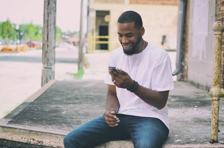 A person is sitting on a concrete platform near a building, casually dressed in a white t-shirt and jeans. They are smiling while looking at a smartphone, indicating engagement and a positive mood. The urban background includes blurred outdoor elements like trees and buildings.