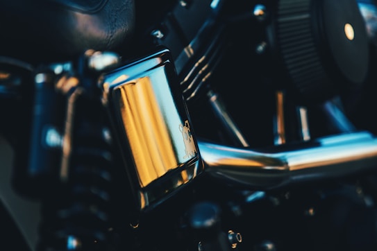 Close-up view of a motorcycle engine with metallic and reflective surfaces, showcasing the intricate design and engineering. The focus is on a chrome component with yellowish reflections, surrounded by various engine parts.
