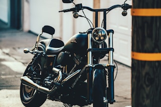 A sleek black motorcycle is parked in an urban setting, positioned at an angle allowing a clear view of its shiny chrome details and front headlamp. The background includes a blurred urban environment, with a wooden utility pole partially obscuring the right side of the image.