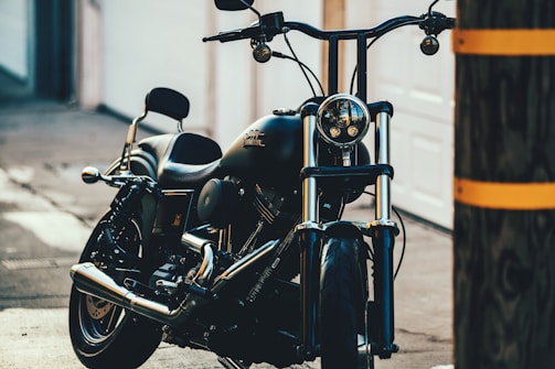 A sleek black motorcycle is parked in an urban setting, positioned at an angle allowing a clear view of its shiny chrome details and front headlamp. The background includes a blurred urban environment, with a wooden utility pole partially obscuring the right side of the image.
