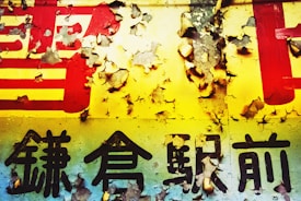 A weathered sign with pealing paint displaying bold, black kanji characters on a colorful background. The paint appears cracked and fragmented, revealing layers beneath.