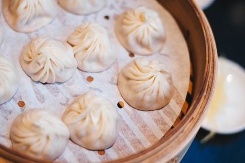 A bamboo steamer basket filled with several neatly arranged dumplings, resting on a perforated sheet. The dumplings have a pleated top and a light, fluffy appearance.