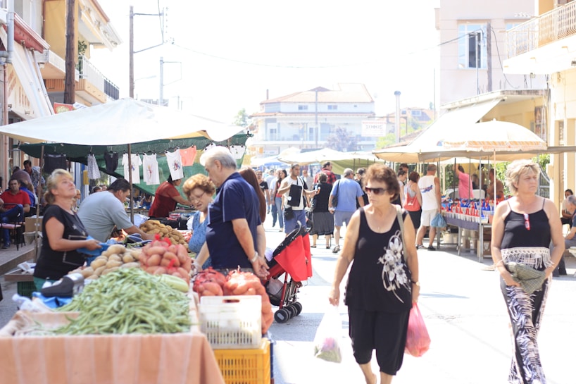 A bustling outdoor market scene with numerous people shopping for fresh produce. Stalls are set up along the street, covered with umbrellas to provide shade, and are filled with various fruits and vegetables like potatoes, tomatoes, and green beans. Shoppers are walking and examining the items on sale, carrying reusable bags.