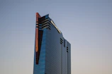 A modern skyscraper with a sleek, reflective glass facade stands tall against a clear sky. The building features dark tinted windows and a series of angular architectural elements, including red-tinted accents along its edges.