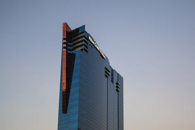 A modern skyscraper with a sleek, reflective glass facade stands tall against a clear sky. The building features dark tinted windows and a series of angular architectural elements, including red-tinted accents along its edges.