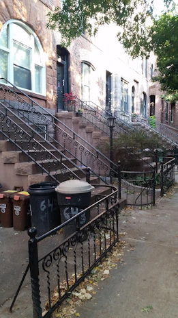 A series of brownstone buildings with ornate iron railings and a set of steps leading up to black doors. Several garbage bins are lined up along the sidewalk, surrounded by fallen leaves. Overhead, some green trees cast partial shadows on the building façade.