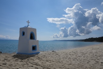 A serene beach scene with a small white and blue chapel-like structure featuring a cross on top, situated on sandy shores beside calm, clear blue water. Large, fluffy clouds fill the sky, adding texture to the bright blue background.