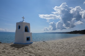 A serene beach scene with a small white and blue chapel-like structure featuring a cross on top, situated on sandy shores beside calm, clear blue water. Large, fluffy clouds fill the sky, adding texture to the bright blue background.