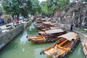 Wooden boats are docked along a narrow, tree-lined canal. The water is a murky green, and stone pathways run parallel on both sides. People are walking along the pathways, some standing and observing the scenery. Overhead, lush trees provide shade, creating a peaceful and serene atmosphere.