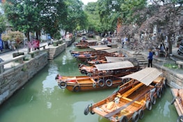 Wooden boats are docked along a narrow, tree-lined canal. The water is a murky green, and stone pathways run parallel on both sides. People are walking along the pathways, some standing and observing the scenery. Overhead, lush trees provide shade, creating a peaceful and serene atmosphere.