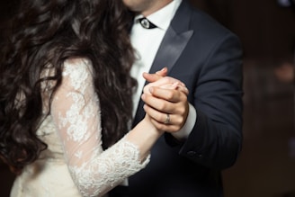 A couple is elegantly dressed, with the man in a dark suit and the woman in a lace dress, sharing a close dance. Their hands are intertwined, conveying a sense of intimacy and romance.