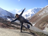 A person wearing outdoor gear poses playfully on a mountain trail with snow-covered peaks and clear blue skies in the background. The landscape features rugged terrain with patches of snow and brown vegetation.