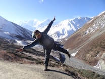 A person wearing outdoor gear poses playfully on a mountain trail with snow-covered peaks and clear blue skies in the background. The landscape features rugged terrain with patches of snow and brown vegetation.
