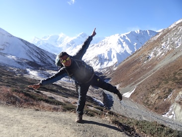 A person wearing outdoor gear poses playfully on a mountain trail with snow-covered peaks and clear blue skies in the background. The landscape features rugged terrain with patches of snow and brown vegetation.
