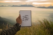 Author writing outdoors with a scenic mountain view and a journal on their lap.