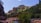 A scenic landscape featuring a rugged canyon with towering red cliffs and lush greenery. A stream runs through the middle with people standing and enjoying the water. The sky is clear with a deep blue hue, and sunlight brightens the canyon walls.