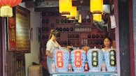 A traditional shop scene featuring two people behind a counter with large, ornate dispensers covered in blue and white patterns. The interior is warmly lit with hanging lanterns and shelves filled with bottles and jars. Various signs with Chinese characters adorn the walls, adding a cultural context.