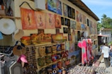 A vibrant outdoor market stall displays a variety of colorful paintings and crafts on the walls. The artwork includes vivid landscapes, abstract figures, and traditional themes. Below the paintings, handmade items such as woven bags, hats, and patterned clothing are on display. Visitors, including a couple of children, are interacting with the stall, exploring the items for sale. The setting appears to be sunny and warm, with a few palm trees visible in the background.