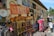 A vibrant outdoor market stall displays a variety of colorful paintings and crafts on the walls. The artwork includes vivid landscapes, abstract figures, and traditional themes. Below the paintings, handmade items such as woven bags, hats, and patterned clothing are on display. Visitors, including a couple of children, are interacting with the stall, exploring the items for sale. The setting appears to be sunny and warm, with a few palm trees visible in the background.