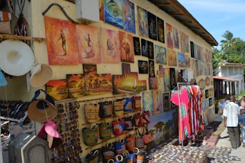 A vibrant outdoor market stall displays a variety of colorful paintings and crafts on the walls. The artwork includes vivid landscapes, abstract figures, and traditional themes. Below the paintings, handmade items such as woven bags, hats, and patterned clothing are on display. Visitors, including a couple of children, are interacting with the stall, exploring the items for sale. The setting appears to be sunny and warm, with a few palm trees visible in the background.