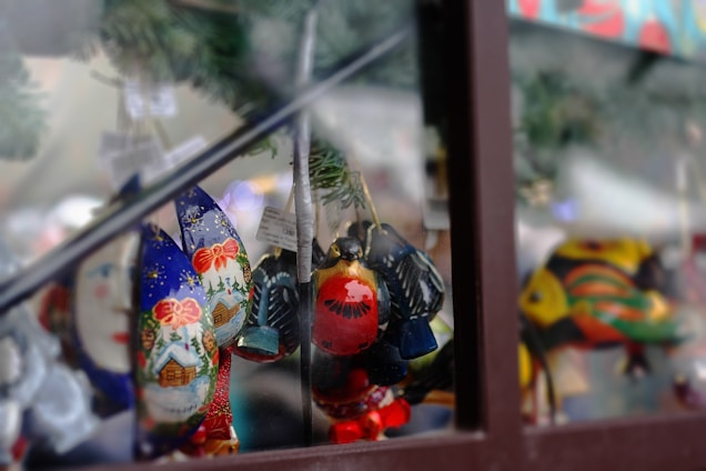 A collection of brightly colored, intricate handcrafted ornaments hangs against a blurred festive background. These decorations feature vibrant designs, including a snow-covered cabin, flowers, and abstract patterns, reflecting holiday themes. The ornaments are made from a glossy material and are hanging from a wire or string, suggesting a festive display.