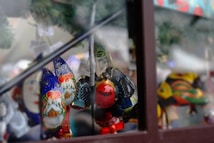 A collection of brightly colored, intricate handcrafted ornaments hangs against a blurred festive background. These decorations feature vibrant designs, including a snow-covered cabin, flowers, and abstract patterns, reflecting holiday themes. The ornaments are made from a glossy material and are hanging from a wire or string, suggesting a festive display.