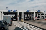 A border checkpoint features several lanes and a white structure with multiple signs indicating passport control. Vehicles, such as cars and a bus, are visible, and there are people both inside and outside the cars. The setting is outdoors under a cloudy sky, and European Union flags are seen above the building.