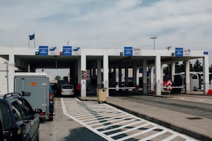 A border checkpoint features several lanes and a white structure with multiple signs indicating passport control. Vehicles, such as cars and a bus, are visible, and there are people both inside and outside the cars. The setting is outdoors under a cloudy sky, and European Union flags are seen above the building.