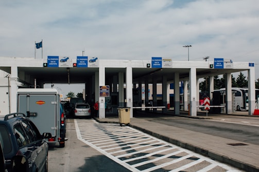 A border checkpoint features several lanes and a white structure with multiple signs indicating passport control. Vehicles, such as cars and a bus, are visible, and there are people both inside and outside the cars. The setting is outdoors under a cloudy sky, and European Union flags are seen above the building.