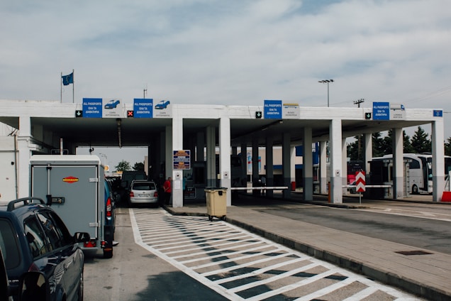 A border checkpoint features several lanes and a white structure with multiple signs indicating passport control. Vehicles, such as cars and a bus, are visible, and there are people both inside and outside the cars. The setting is outdoors under a cloudy sky, and European Union flags are seen above the building.