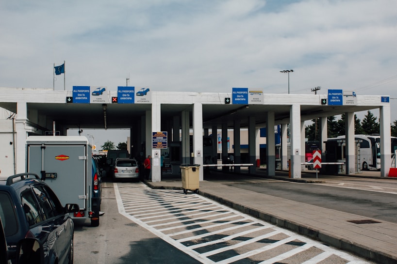 A border checkpoint features several lanes and a white structure with multiple signs indicating passport control. Vehicles, such as cars and a bus, are visible, and there are people both inside and outside the cars. The setting is outdoors under a cloudy sky, and European Union flags are seen above the building.