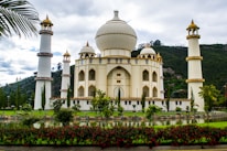 A majestic building with architectural features reminiscent of the Taj Mahal, surrounded by four tall minarets. The structure is cream-colored with gold accents and is set against a backdrop of lush green hills, with a manicured garden and reflective pond in the foreground. Red flowers and tall trees add to the scenic beauty.