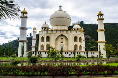 A majestic building with architectural features reminiscent of the Taj Mahal, surrounded by four tall minarets. The structure is cream-colored with gold accents and is set against a backdrop of lush green hills, with a manicured garden and reflective pond in the foreground. Red flowers and tall trees add to the scenic beauty.