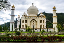 A majestic building with architectural features reminiscent of the Taj Mahal, surrounded by four tall minarets. The structure is cream-colored with gold accents and is set against a backdrop of lush green hills, with a manicured garden and reflective pond in the foreground. Red flowers and tall trees add to the scenic beauty.