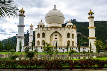 A majestic building with architectural features reminiscent of the Taj Mahal, surrounded by four tall minarets. The structure is cream-colored with gold accents and is set against a backdrop of lush green hills, with a manicured garden and reflective pond in the foreground. Red flowers and tall trees add to the scenic beauty.